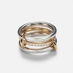May include: A multi-band ring featuring two smooth silver-toned bands and two gold-toned bands. One gold-toned band is set with a row of small, clear, round-cut stones. The bands are interconnected by two small gold-toned loops on the sides.