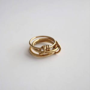 May include: A gold-colored ring with a unique knot design. The ring features multiple bands that intertwine to create a stylish and modern look. The ring is made of a shiny, metallic material and is set against a plain white background.