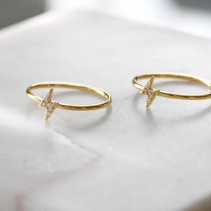 May include: Two gold-colored rings with a lightning bolt design. Each ring features a thin band and a lightning bolt charm encrusted with small, clear gemstones. The rings are displayed on a white surface, creating a bright and elegant aesthetic.