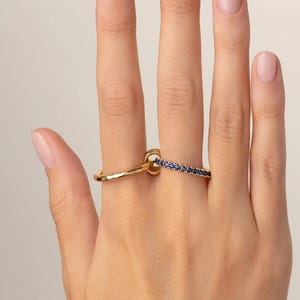 May include: A gold ring with a smooth, undulating band linked to a second gold ring featuring a row of small, round, deep blue gemstones set along the outer edge. The two rings are connected, creating a singular, layered jewelry piece.