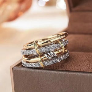 Op de afbeelding: Een multi-band ring met goud- en zilverkleurige tinten. De ring heeft meerdere banden, waarvan sommige zijn bezet met kleine, heldere edelstenen. De banden zijn verbonden met goudkleurige accenten en een zilverkleurig knoopontwerp.