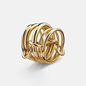 May include: A gold-colored ring with a complex, interwoven design. The ring features multiple rounded bands that are interlocked, creating a unique and eye-catching appearance. The metal has a polished, reflective surface.