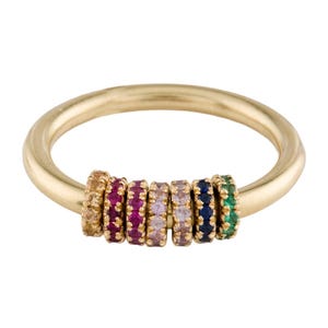 Op de afbeelding: Een gouden ring met zeven kleine, ronde, met edelstenen bezette bedels die langs de ringband schuiven. De stenen zijn in een rij geplaatst en hebben de kleuren geel, magenta, donkerroze, lichtroze, lavendel, donkerblauw en heldergroen.