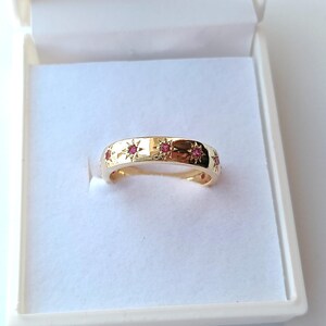 May include: A gold band ring featuring a series of small, round, pink gemstones set within engraved starburst patterns that are evenly spaced around the outer surface of the polished metal band, presented in a white jewelry box.