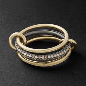 May include: A multi-band ring featuring two smooth yellow gold bands and one dark metal band. The central dark band is encrusted with a pavé of small, alternating brown and white round-cut stones, creating a textured, sparkling appearance.