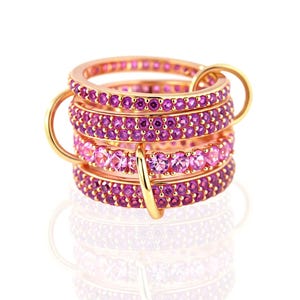 Op de afbeelding: Stapel goudkleurige ringen met roze en paarse edelstenen. De ringen zijn verbonden door gouden lussen, waardoor een gelaagd effect ontstaat. De edelstenen zijn in rijen gerangschikt, met een weerspiegeling van de ringen eronder.