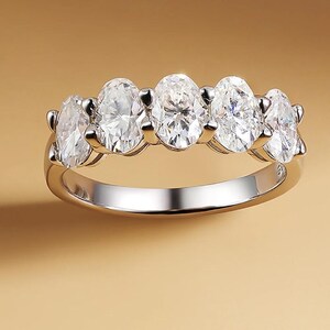 Könnte beinhalten: Ein silberner Ring mit fünf oval geschliffenen, klaren Edelsteinen. Die Edelsteine sind in einer Reihe über der Oberseite des Rings angeordnet, mit einem polierten Silberband. Der Ring wird vor einem warmen, neutralen Hintergrund präsentiert.