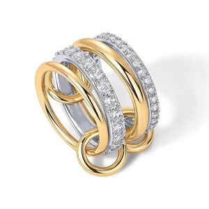 Puede incluir: Un anillo multibanda en tonos dorados y plateados. Una banda está incrustada con pequeñas piedras preciosas transparentes. El anillo presenta bucles entrelazados y un anillo dorado colgante. El diseño es moderno y elegante.