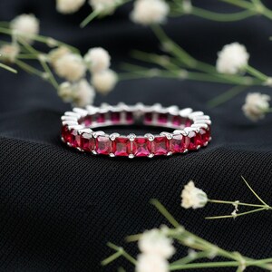Könnte beinhalten: Ein silberner Eternity-Ring mit einer durchgehenden Reihe quadratisch geschliffener, leuchtend roter Edelsteine in einer polierten Metallfassung. Der Ring hat ein einheitliches Design, bei dem jeder Stein von kleinen Metallkrappen gehalten wird.