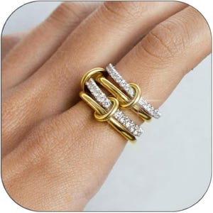 May include: Two gold rings with a knot design, each featuring a row of small, sparkling white stones. The rings are displayed on a finger, showcasing their intricate design and elegant appearance. The rings have a polished, shiny finish.