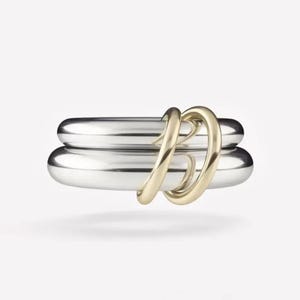 May include: Two silver-coloured, rounded rings stacked together, with a gold-coloured ring intertwined around them. The gold ring has a curved, abstract design. The background is plain white.