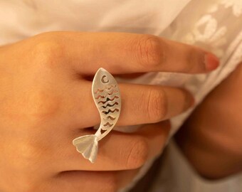 May include: A silver fish-shaped ring. The ring has a detailed design with cut-out wave patterns along the body and a fin at the tail. The fish ring is worn on a finger.