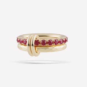 Op de afbeelding: Een gouden ringenset bestaande uit twee in elkaar grijpende ringen. Eén ring is voorzien van een rij ronde, felrode edelstenen in gouden zettingen. De tweede ring heeft een getextureerde, gehamerde afwerking. De twee ringen zijn verbonden door een gladde gouden lus.