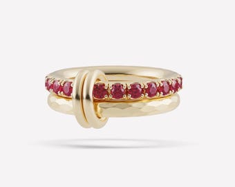 Può includere: Un set di anelli in oro composto da due fasce intrecciate. Una fascia presenta una fila di pietre preziose rosse rotonde incastonate in oro. La seconda fascia ha una finitura martellata. I due anelli sono uniti da un anello d'oro liscio.