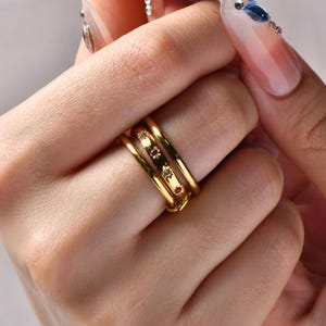 May include: A set of four gold rings, stacked on a finger. The rings have a smooth, polished finish. Three of the rings are adorned with small, sparkling gemstones in red and clear colors. The rings are a classic and elegant jewelry item.