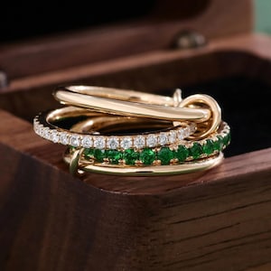 May include: A stack of gold rings with different designs. One ring features a row of small, sparkling diamonds, while another has a row of vibrant green gemstones. The rings are displayed inside a wooden box.