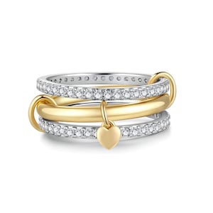 May include: A multi-band ring featuring two silver-toned bands encrusted with small clear stones and a central smooth gold-toned band. The bands are linked together by a gold-toned loop, and a small, solid gold-toned heart charm hangs from the center.