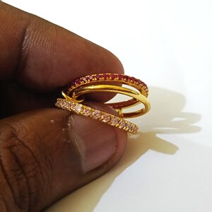 May include: A set of three interlocking rings featuring one gold band, one band encrusted with small red stones, and one band encrusted with small light pink stones. The rings are connected to form a single cohesive piece of jewellery.