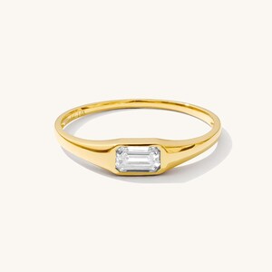 Puede incluir: Un anillo de oro con una piedra preciosa rectangular y transparente. El anillo tiene una banda lisa y pulida y un engaste de bisel para la piedra preciosa. El anillo está sobre un fondo blanco.