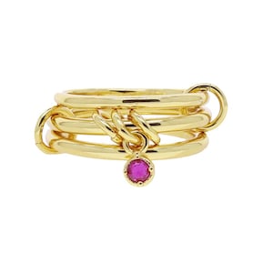 May include: Gold-toned ring with multiple linked bands. A small, round, magenta-colored gemstone hangs from the center. The ring has a polished finish and a unique, layered design. The ring is a piece of jewelry.
