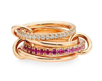 Può includere: Un insieme di quattro anelli d'oro collegati tra loro. Uno è incastonato con piccoli diamanti bianchi, un altro con pietre preziose rosa. Gli anelli sono in oro lucido e collegati da grandi maglie arrotondate.