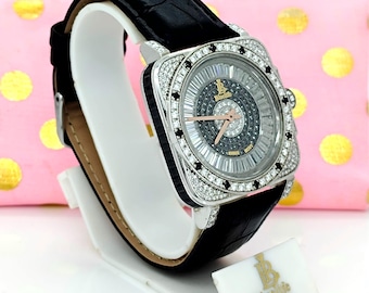 May include: A square-faced wristwatch with a black leather band. The watch features a silver-colored bezel encrusted with clear and black gemstones. The watch face is silver and black with a radial design and a small logo. The watch is displayed on a white stand.