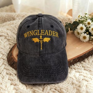 Może przedstawiać: Ciemnoszara czapka bejsbolowa z napisem "WINGLEADER" wyhaftowanym złotą nicią nad złotym emblematem. Czapka ma zakrzywiony daszek i klasyczny sześciopanelowy design. Kapelusz leży na kremowej powierzchni.