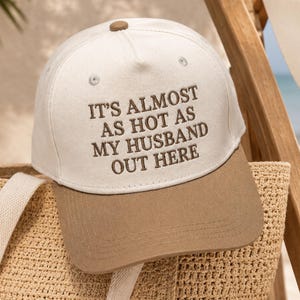 May include: A beige and brown baseball cap with the embroidered text "IT'S ALMOST AS HOT AS MY HUSBAND OUT HERE." The cap is resting on a woven tote bag, suggesting a beach or outdoor setting.