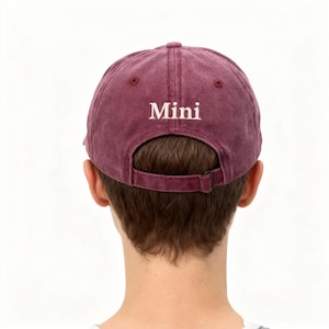 May include: A maroon baseball cap with a distressed, washed-out texture. The word Mini is embroidered in white, serif font above the back opening. The cap features a matching maroon fabric strap with a metal buckle for an adjustable fit.