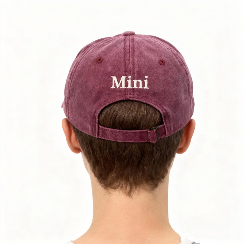 May include: A maroon baseball cap with a distressed, washed-out texture. The word Mini is embroidered in white, serif font above the back opening. The cap features a matching maroon fabric strap with a metal buckle for an adjustable fit.