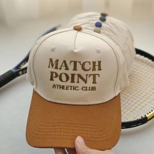 May include: A beige and brown baseball cap with the text "MATCH POINT ATHLETIC CLUB" embroidered on the front. The cap has a curved brim and a button on top. A tennis racket is in the background.