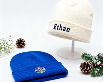 May include: Two knit beanies. One is cream-colored with the name "Ethan" embroidered in navy blue. The other is bright blue with a colorful embroidered patch. The beanies are displayed on a white surface with decorative pine cones and greenery.
