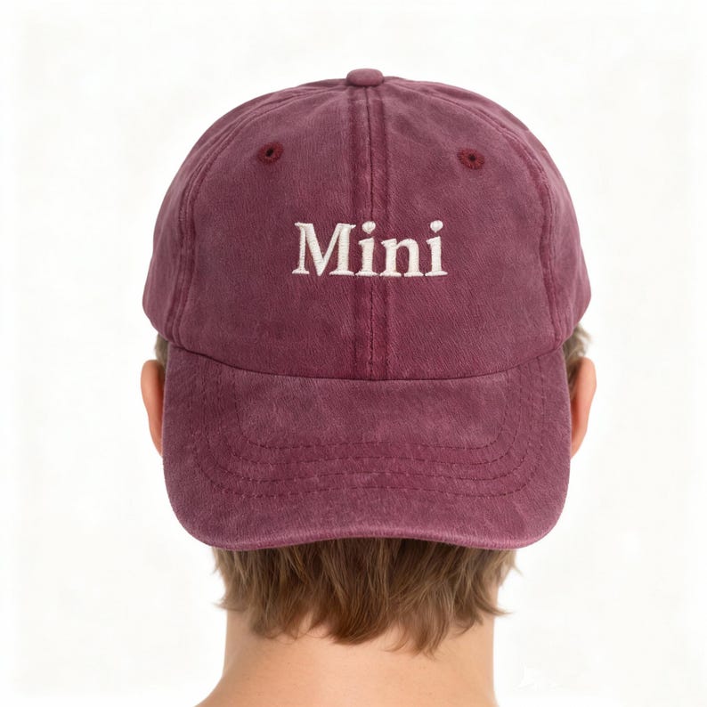 May include: A maroon baseball cap with a six-panel construction and a curved brim. The word "Mini" is embroidered in white, serif-style lettering across the front center of the cap. The fabric has a slightly faded, washed-out appearance.