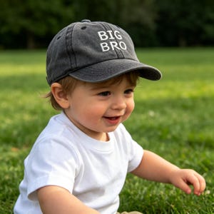 May include: A charcoal gray baseball cap with a distressed, washed-out finish. The front panel features the text BIG BRO embroidered in bold, white, sans-serif capital letters. The cap has a curved brim and a structured crown design.