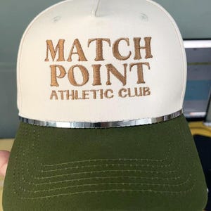 May include: A white and olive green baseball cap with the text "MATCH POINT ATHLETIC CLUB" embroidered in brown. The cap has a silver trim and a green button on top.