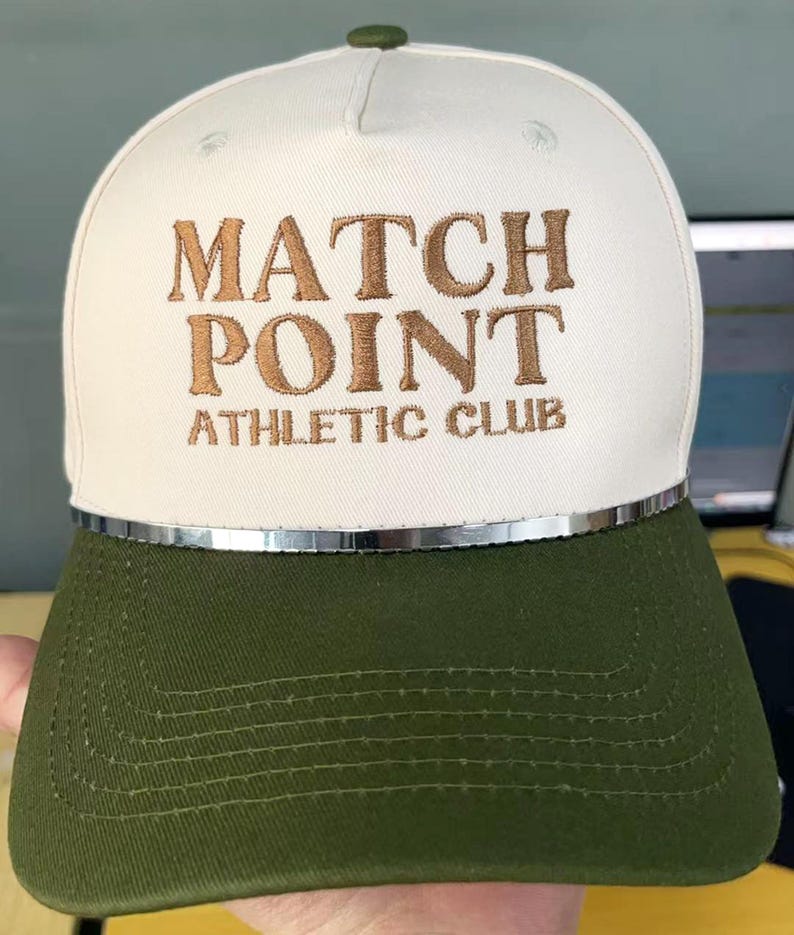 May include: A white and olive green baseball cap with the text "MATCH POINT ATHLETIC CLUB" embroidered in brown. The cap has a silver trim and a green button on top.