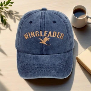 Peut inclure: Casquette de baseball bleue avec le mot "WINGLEADER" brodé en fil d'or. Un motif d'oiseau doré est en dessous du texte. La casquette a une visière incurvée et un bouton sur le dessus. Le chapeau est posé sur une table.