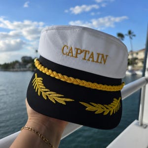 Może przedstawiać: Biało-czarna czapka kapitana ze złotym haftowanym tekstem i akcentami. Słowo "CAPTAIN" jest wyhaftowane złotem na białej górze. Złota lina i złoty wzór liści zdobią rondo kapelusza, sugerując motyw marynistyczny.