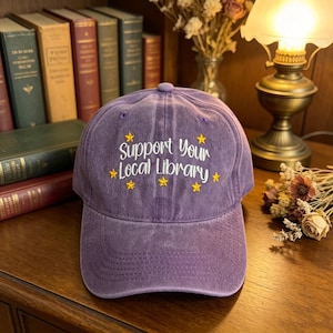 Peut inclure: Casquette de baseball violette avec l'inscription brodée "Support Your Local Library" en blanc, ornée d'étoiles jaunes. La casquette a une visière incurvée et une conception classique à six panneaux.