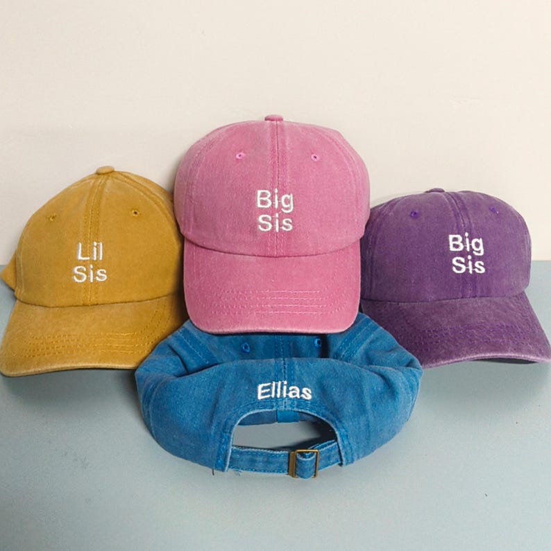 May include: Four baseball caps in mustard yellow, pink, purple, and blue. The yellow cap features white embroidered text reading Lil Sis. The pink and purple caps feature white embroidered text reading Big Sis. The blue cap features white text reading Ellias.