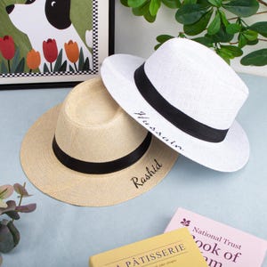 May include: Two fedora hats, one white and one beige, with black bands. The white hat has the name "Hussain" embroidered on it, and the beige hat has "Rashid" embroidered. The hats are on a light blue surface.