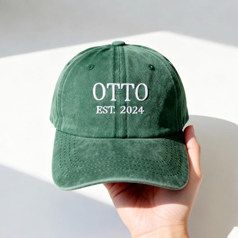 May include: A forest green baseball cap with a washed, textured finish. The front features white embroidered text that reads OTTO in a serif font, with the text EST. 2024 embroidered in a smaller white serif font directly underneath.