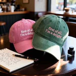 May include: Two baseball caps, one pink and one green, with the embroidered text "first of all, I'm a delight." The caps are sitting on a dark wooden table next to a notebook and pen.