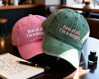 May include: Two baseball caps, one pink and one green, with the embroidered text "first of all, I'm a delight." The caps are sitting on a dark wooden table next to a notebook and pen.
