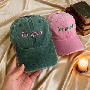 以下が含まれることがあります： 「for good」の刺繍が入った、濃い緑色とピンク色のベースボールキャップが2つ。キャップは柔らかく、質感のある生地で作られています。緑色のキャップにはピンク色の刺繍、ピンク色のキャップには緑色の刺繍が施されています。