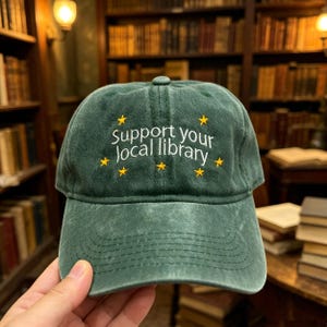 May include: Green baseball cap with the text "Support your local library" in white and yellow star accents. The cap has a curved brim and a classic six-panel design. The hat is made of a soft, worn material.