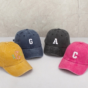 Könnte beinhalten: Vier Baseballkappen in verschiedenen Farben: Senfgelb, Marineblau, Anthrazitgrau und Pink. Jede Kappe hat einen weißen, gestickten Buchstaben oder ein Monogramm. Die Kappen sind auf einer weißen Oberfläche angeordnet.