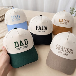 Puede incluir: Cinco gorras de béisbol con coronas blancas y viseras de colores. Cada gorra tiene texto bordado: "DAD 06.15.2026", "PAPA EST. 2026", "DADDY SINCE 2026", "DAD EST. 2026" y "GRANDPA EST. 2026". Las viseras son azules, negras, marrones, verdes y grises.