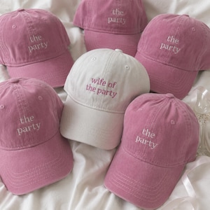 May include: A collection of six baseball caps made of cotton fabric. Five caps are dusty rose pink with the text "the party" embroidered in white thread. One central cap is off-white with the text "wife of the party" embroidered in pink.