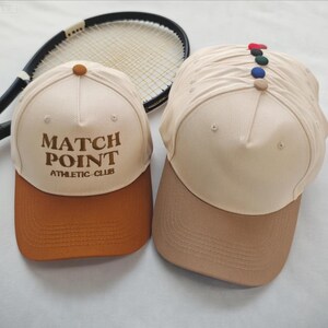 May include: Two baseball caps with curved brims. One is tan with a brown brim and the text "MATCH POINT ATHLETIC CLUB" in brown. The other is tan with a brown brim and a row of colorful buttons on top. A tennis racket is in the background.